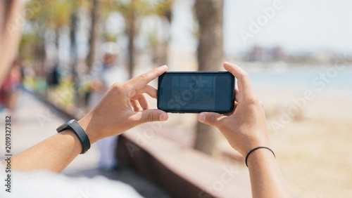 Person takes a picture on a phone while walking near the beach on a sunny day with palm trees in view
