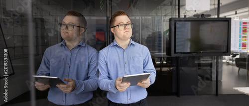 Professional man, corporate staff member with Down syndrome stands next to glass blackboard with digital tablet in office, looks away with pensive expression, thinks on ideas, planning, strategizing