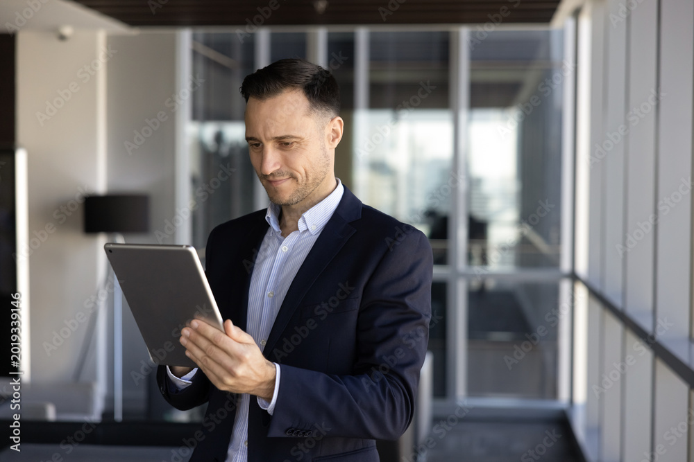 Fototapeta premium Focused businessman stands in office hallway with tablet, managing business tasks, reads information, review digital data, consulting corporate client via email, staying connected through technology