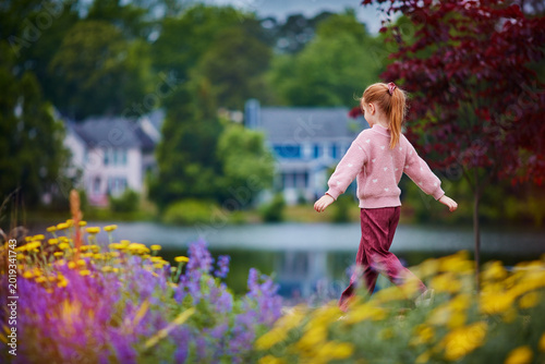 Dreamy Summer Childhood Scene with Little Girl Walking Through Blooming Flower Field in Peaceful Neighborhood