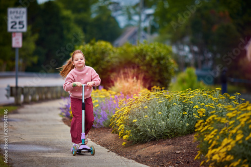 Active Little Girl Enjoying Outdoor Scooter Ride Along Flower-Lined Sidewalk in Summer Suburban Neighborhood