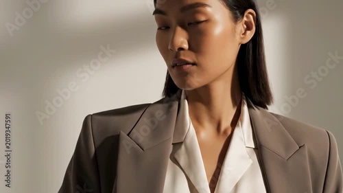 Sophisticated asian model in oversized taupe blazer and white blouse, sharp sunlight, clean beige wall, poised business fashion expression, and premium editorial styling for modern leadership image