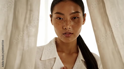 Freckled asian model portrait in white linen shirt between soft curtains, glowing backlight, centered gaze, clean natural beauty mood, and elegant lifestyle fashion styling for premium soft image