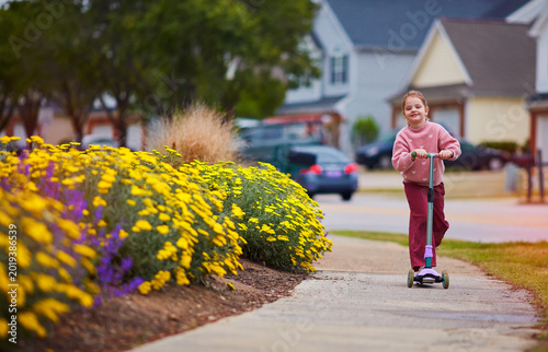 Happy Girl Enjoying Outdoor Scooter Ride Along Flower-Lined Sidewalk in Summer Suburban Neighborhood