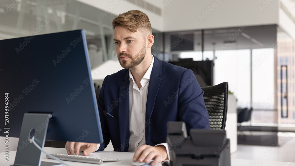 Fototapeta premium Young man, office employee sits at desk, read emails or important correspondence, writing or editing proposal and document, evaluating financial or project planning information, strategizing using PC