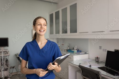 Φωτογραφία Portrait of smiling female nurse in blue medical scrubs holding clipboard stands