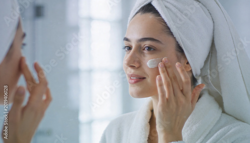 Woman wrapped in towel applying moisturizer in bright bathroom