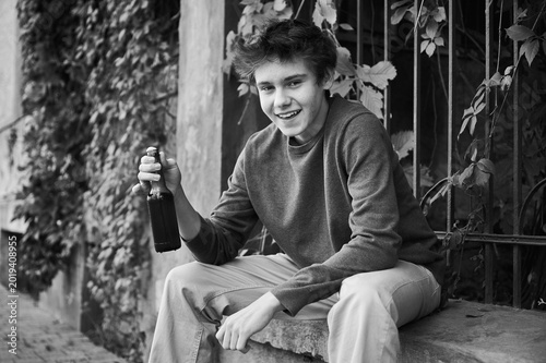 Teen male drinks alone from bottle on sidewalk. Emotional urban scene presents vulnerable adolescent with troubled mood and isolated posture. Concept of drinking problem among youth