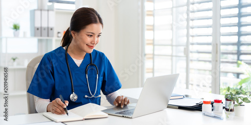 Obraz na plátně Doctor looking at laptop screen writing in notebook for research planning or medical tech innovation in hospital office