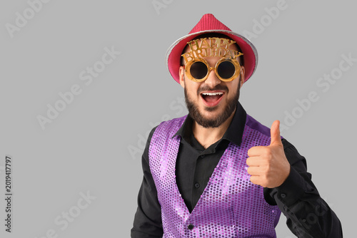 Cheerful young man with HAPPY BIRTHDAY party glasses showing thumb-up on grey...