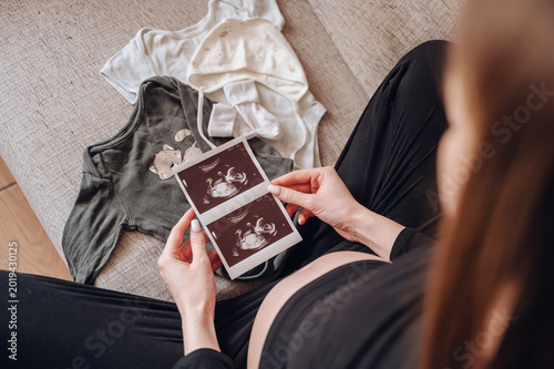 Close up of a pregnant woman holding ultrasound images of her baby next to tiny clothes on a sofa