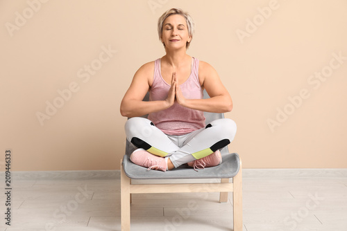 Portrait of meditating mature woman sitting on chair against beige wall