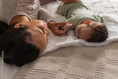Obraz na płótnie Mother holding infant's hand lying on bed on plush blanket, beige throw, wearing