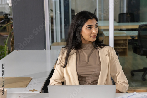 Indian woman sitting at office desk wearing beige blazer using laptop, document, mug, pad