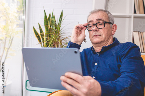 elderly man has difficulty reading a document on a digital tablet while wearing glasses.
