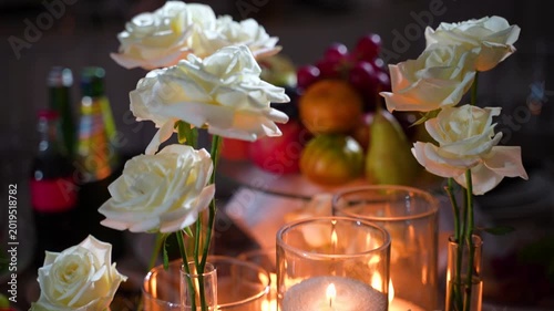 Restaurant table decorated with white roses, featuring platters of food and fruit. Red apples and candles with flowers.