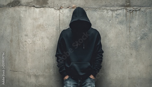 Person in dark hoodie, face obscured, hands in pockets, against textured wall