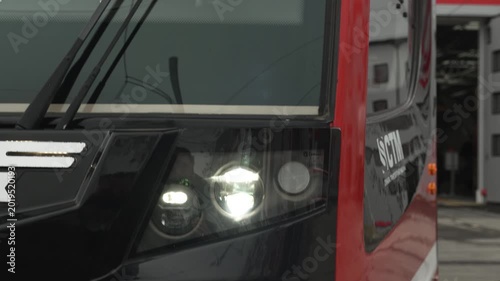 New red tram with its headlights on leaves the depot during the day. Modern, electric, environmentally friendly urban transport. Bright light from headlight shining into the camera from train car