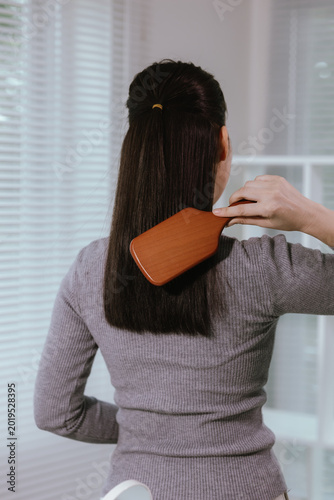 Back view of a young Asian woman brushing her long straight hair at home, showing healthy hair care and daily beauty routine.