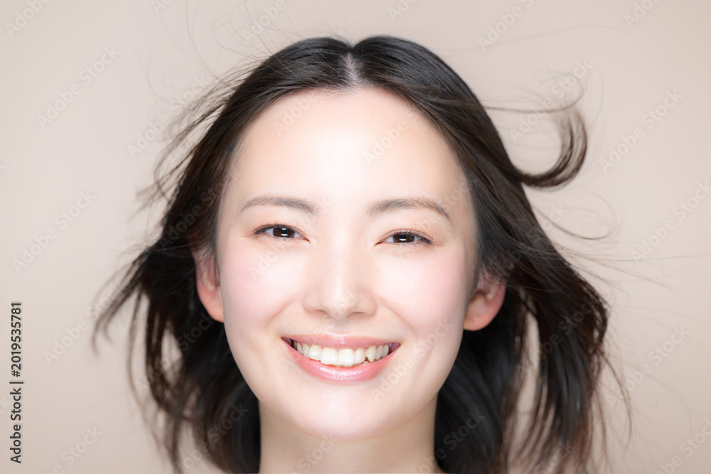 Naklejka premium ベージュ背景の風になびくサラサラ黒髪と笑顔の女性美肌と透明感あるスキンケア美容やトリートメントイメージクローズアップカメラ目線