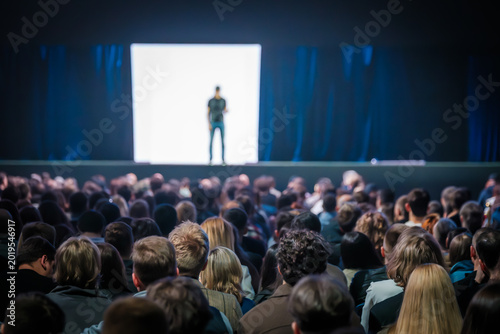 Large audience watching speaker on stage during conference presentation, bright screen backdrop and focused spotlight creating dramatic atmosphere.
