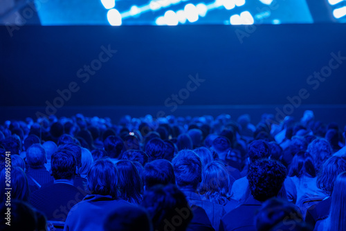 Bild auf Leinwand Large audience watching presentation in dark blue conference hall, corporate event atmosphere, focused attention and anticipation