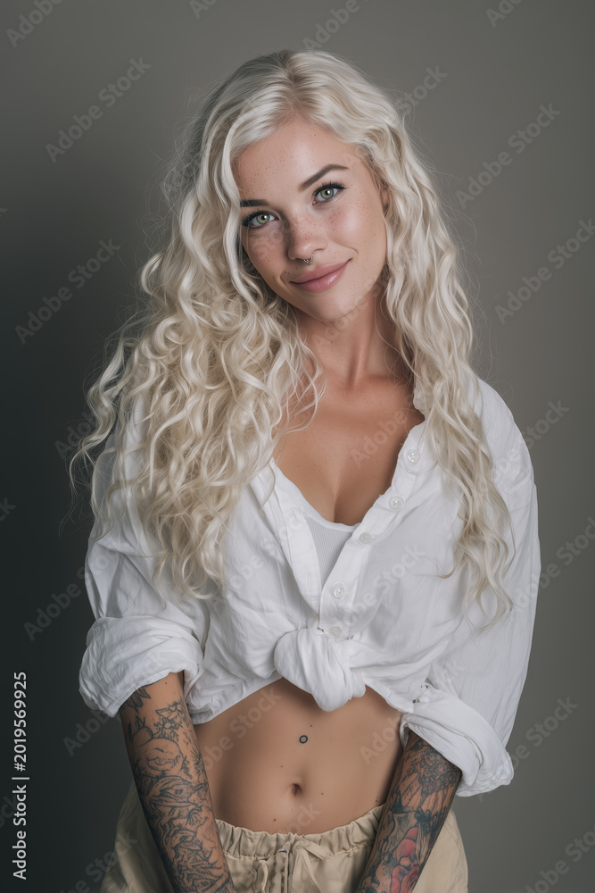Naklejka premium Young woman with long curly blonde hair and tattoos wearing a white shirt and light pants poses with a smile against a neutral background