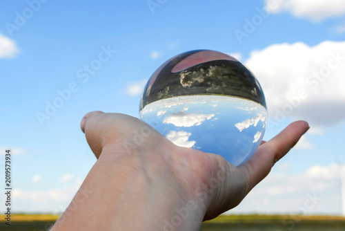 Creative photography of blue sky and white clouds refracted inside a glass sphere held by a human hand. Optical illusion and clean environment concept.