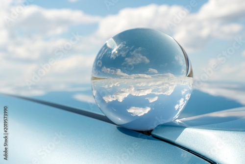 Crystal lensball refracting clean sky and horizon on a blue car hood. Creative photography representing clean energy, future vision, and environment.