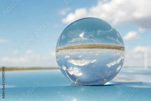 Minimalist concept with a glass sphere reflecting blue sky and clouds, resting on a sleek, glossy blue metallic surface. Futuristic abstract background.
