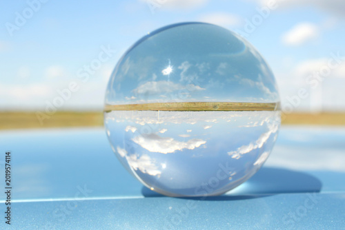 Minimalist concept with a glass sphere reflecting blue sky and clouds, resting on a sleek, glossy blue metallic surface. Futuristic abstract background.