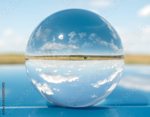 Minimalist concept with a glass sphere reflecting blue sky and clouds, resting on a sleek, glossy blue metallic surface. Futuristic abstract background.
