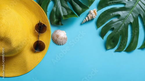 Yellow straw hat with sunglasses, seashell, and green monstera leaf on bright blue background, flat lay top view summer banner with copy space