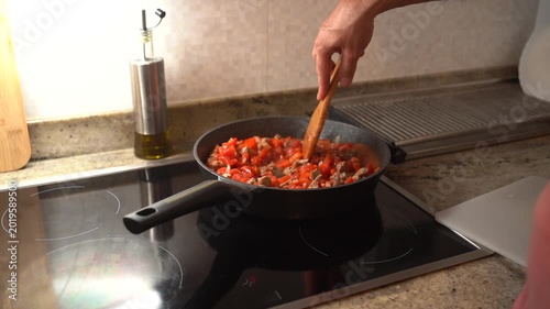 Close-up of a frying pan with beef, onion, and diced red sweet pepper being cooked on a stove in a home kitchen.
