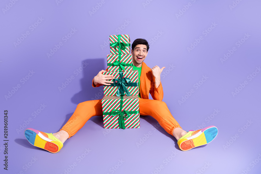 Naklejka premium Happy man in bright orange suit holding stacked Christmas gifts on purple background with colorful sneakers for a playful festive celebration