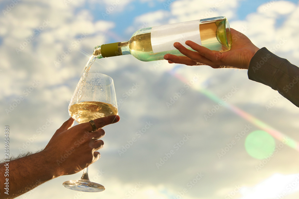 Fototapeta premium White wine pouring into glass against bright sky with sun flare. Concept of premium winery promo, summer aperitif, outdoor celebration, restaurant menu and luxury travel.