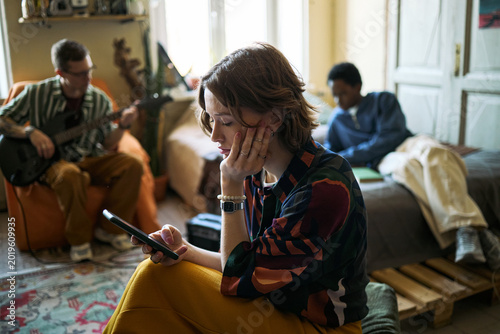 Quadro em tela Young adult Caucasian woman checking smartphone while sitting with friends at home, showing digital distraction and social disconnect