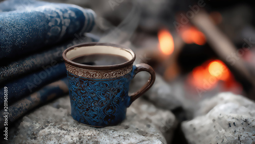 Steaming mug of dark coffee rests on rocks beside a cozy campfire and blankets