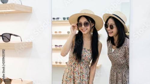 Two girls in straw hats admire their reflection while trying on sunglasses. The image feels bright and stylish, capturing a moment of genuine connection and trendy summer fun.