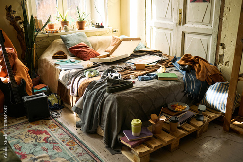 Fotografi Wide angle shot of messy young adult bedroom showing pizza boxes, books, headphones, guitar, takeaway coffee, suggesting student burnout or chaotic remote living