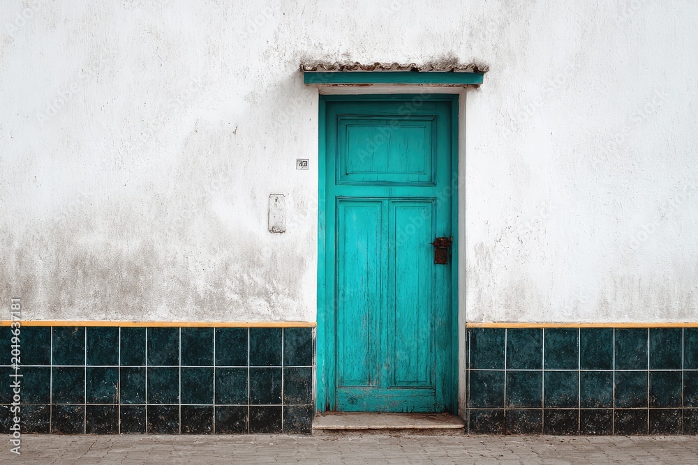 Fototapeta premium Weathered blue door centered on a rough white wall, bordered by tile