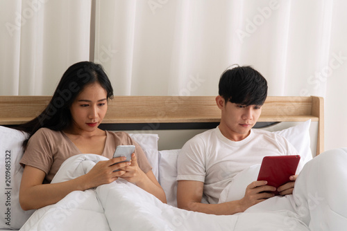 Young Asian couple lying in bed ignoring each other while using their own smart devices. Man holding a tablet and woman looking at smartphone. Relationship problem and digital addiction concept.