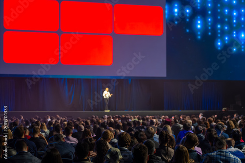 Conference speaker on stage addressing large audience during business presentation, bright screen visuals, professional event atmosphere.
