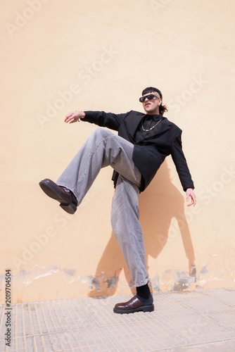 A young man with one leg raised in a dynamic pose in front of a peach-colored outdoor wall