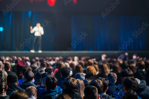 Large audience watching speaker on stage during conference, blurred presenter, rows of attendees focused on keynote talk.