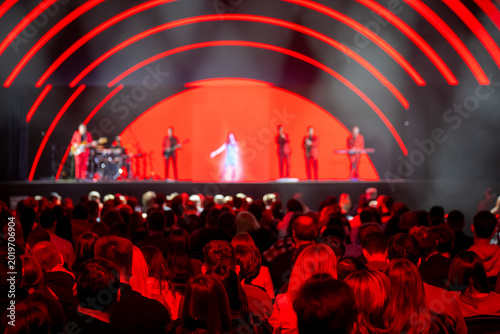 Live concert, band on red illuminated stage, crowded audience watching performance, energetic atmosphere, dramatic stage lights.