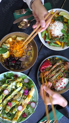 Friends enjoying a vibrant and healthy lunch together at a restaurant, eating delicious asian fusion dishes like ramen, curry, and fresh salads with chopsticks from a dark wooden table