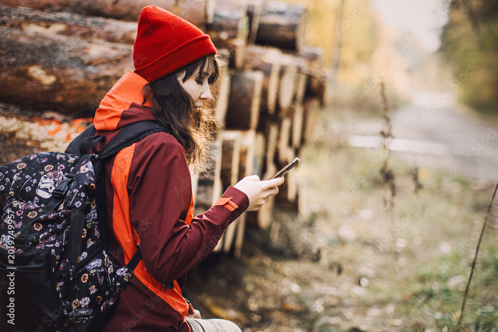 Naklejka premium Woman uses smartphone while resting near logs in autumn forest . Responsible travel destinations, low-impact tourism, local forest exploration.