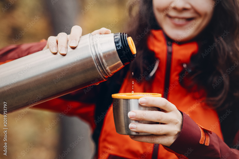 Fototapeta premium Smiling woman pours hot drink from metal thermos into cup outdoors. vacuum insulation technology, thermal efficiency engineering, functional product feature.