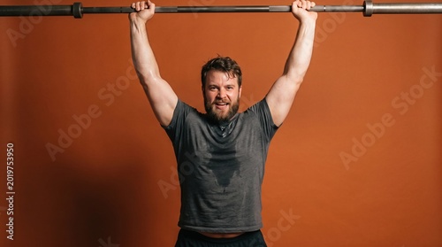 Tapety artystyczne A muscular Caucasian man with a beard lifts a barbell overhead in a gym setting. He wears a gray t-shirt and appears focused and determined.